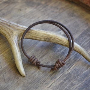 May include: A brown leather bracelet with two knots. The bracelet is on a wooden surface with a piece of antler.