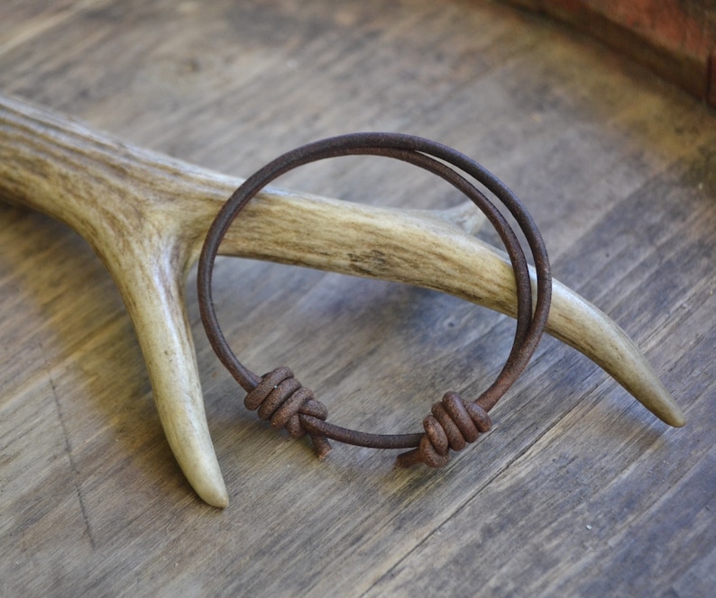 May include: A brown leather bracelet with two knots. The bracelet is on a wooden surface with a piece of antler.