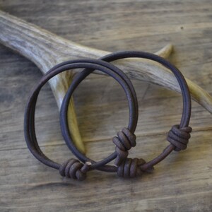 May include: Two brown leather bracelets with knotted closures. The bracelets are lying on a wooden surface.