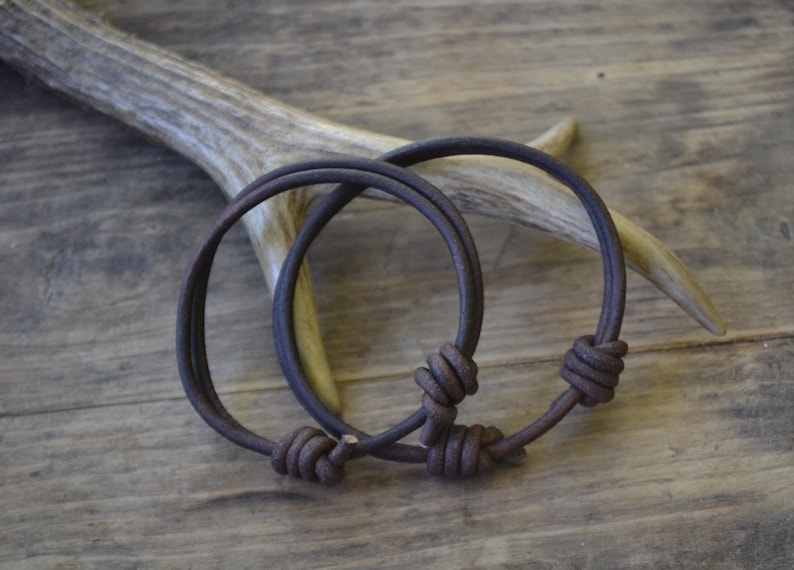 May include: Two brown leather bracelets with knotted closures. The bracelets are lying on a wooden surface.
