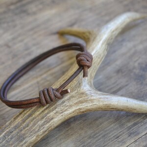May include: A brown leather bracelet with a knotted design, displayed on a white antler.