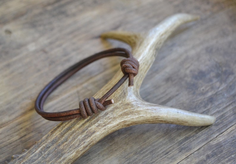 May include: A brown leather bracelet with a knotted design, displayed on a white antler.