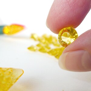 May include: A close-up of a yellow, faceted, round, glass gem held between two fingers. The gem is clear and has a slight sparkle.