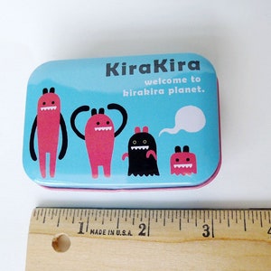 May include: A blue tin with a pink rim featuring the text "KiraKira welcome to kirakira planet." and four cartoon monsters. The monsters are red, black, and pink.