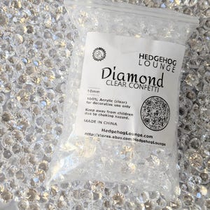 May include: A clear plastic bag of clear diamond confetti from Hedgehog Lounge. The bag contains 10mm, 100% acrylic confetti, for decorative use. The bag has a QR code and the text "Diamond Clear Confetti".