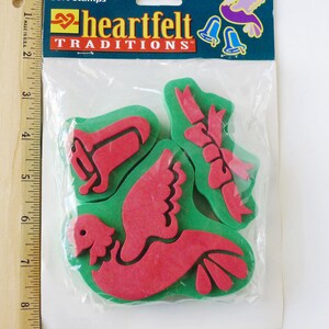 May include: A set of four green and red foam stamps featuring a dove, a bell, and two bows. The stamps are packaged in a clear plastic bag with a header that reads "heartfelt traditions".