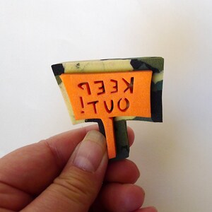 May include: A camouflage-patterned foam sign with an orange cutout that reads "KEEP OUT!"