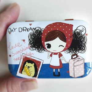 May include: A white and blue compact with a cartoon girl wearing a red and white polka dot hooded dress. The text "DAY DREAM" is printed on the top of the compact. The girl is holding a pink polka dot suitcase. The text "love's sunshine melody" is printed on the side of the compact.