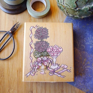 May include: A wooden stamp with a pink and green illustration of a potted plant with a bowl of pink hearts. The plant has a pink bow and ribbon. The stamp is on a wooden surface.