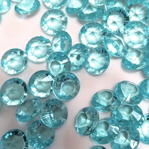 May include: A scattering of turquoise blue, faceted, round, plastic gems. The gems are scattered on a white surface.