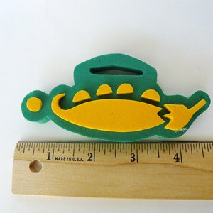 May include: Green and yellow foam shape of a submarine with a yellow circle on the side. The submarine is about 4 inches long.
