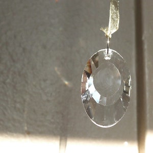 May include: A clear crystal prism pendant hanging from a light beige ribbon. The prism is oval shaped and has a faceted design.