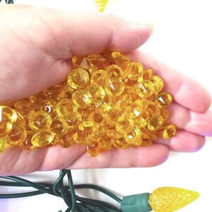 May include: A hand holding a large pile of yellow, faceted, plastic gems. The gems are small and have a diamond-like shape.