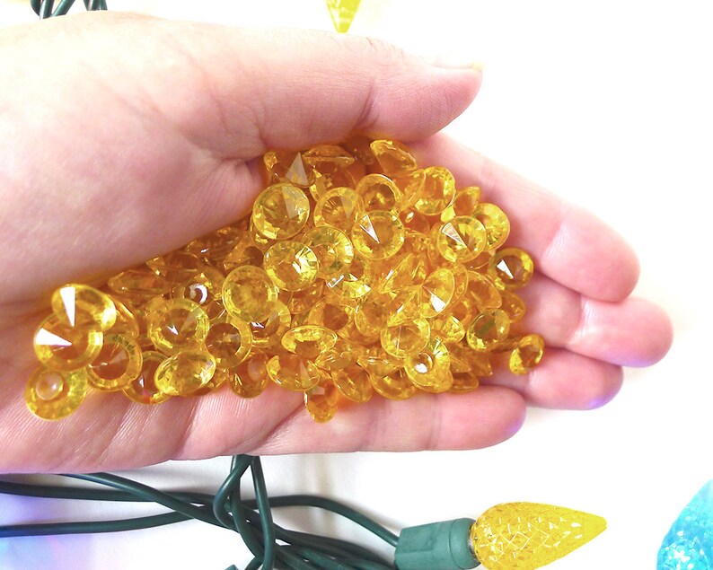 May include: A hand holding a large pile of yellow, faceted, plastic gems. The gems are small and have a diamond-like shape.