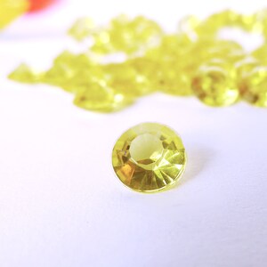 May include: A close-up of a single, clear, yellow, faceted gemstone on a white surface. The gemstone is surrounded by a scattering of similar gemstones.