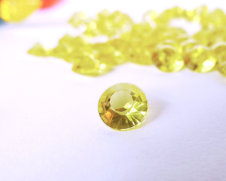 May include: A close-up of a single, clear, yellow, faceted gemstone on a white surface. The gemstone is surrounded by a scattering of similar gemstones.