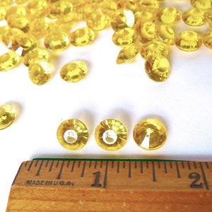 May include: A close-up of yellow diamond-shaped confetti, scattered on a white surface. A ruler is in the foreground, with the numbers "1" and "2" visible.