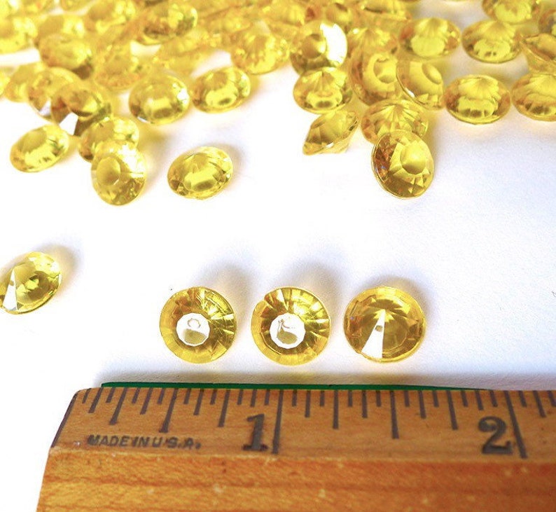 May include: A close-up of yellow diamond-shaped confetti, scattered on a white surface. A ruler is in the foreground, with the numbers "1" and "2" visible.