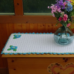 May include: White crocheted table runner with a blue and green floral design. The runner is on a wooden table with a heart design.