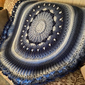 Op de afbeelding: Een handgemaakte, ronde gehaakte deken in tinten blauw en wit. De deken heeft een centraal spiraalontwerp, omgeven door concentrische ringen in verschillende blauwtinten. De randen zijn afgewerkt met een geschulpte rand.