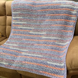 May include: A handmade crochet blanket in shades of lavender, peach, and white, draped over a brown sofa. The blanket features horizontal stripes and a textured stitch pattern, perfect for adding warmth and style to any living space.