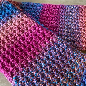 May include: A handmade, textured scarf in shades of blue, pink, and orange. The scarf is made with a thick yarn, creating a woven pattern. The colors blend together, creating a gradient effect. The scarf is folded over itself.