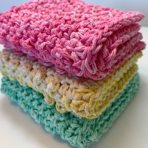 May include: Three crocheted dishcloths stacked on top of each other. The top dishcloth is pink, the middle dishcloth is yellow and white, and the bottom dishcloth is green.