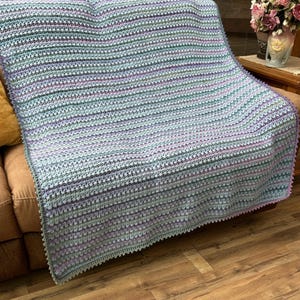 May include: A handmade crochet blanket in shades of blue, green, and purple, draped over a brown sofa. The blanket features a textured stitch pattern and a scalloped edge, creating a cozy and decorative home textile.