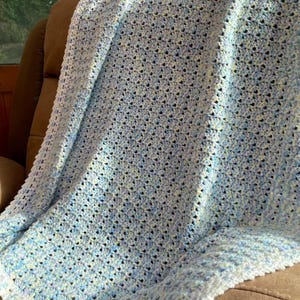 May include: A handmade crochet blanket in shades of light blue, white, and yellow. The blanket is draped over a brown chair, showcasing its textured stitch pattern. The edges have a decorative border, adding a finished look to this cozy, soft blanket.