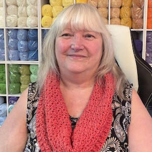 May include: A coral-colored crocheted infinity scarf with a textured stitch pattern. The scarf is worn around the neck over a sleeveless black and white paisley patterned top. The accessory features a consistent yarn weight and loop construction.