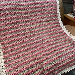 May include: A handmade crochet blanket with a striped pattern in shades of pink, green, and gray. The blanket features a decorative white lace border, adding a delicate touch. This cozy blanket is perfect for adding warmth and style to any home.