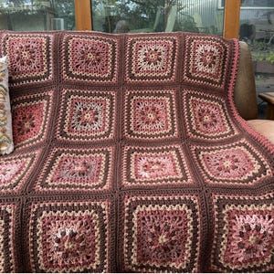 May include: A handmade crochet blanket composed of multiple square motifs in shades of dark brown, dusty rose, and cream. Each square features a central floral-like pattern surrounded by concentric rows of yarn, joined together to form a large throw.