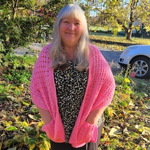 May include: A pink crocheted shawl with pockets. The shawl is open in the front and has a lacy pattern. The person wearing the shawl is standing in front of a background of fall leaves.