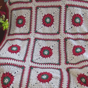 Könnte beinhalten: Eine weiße Häkeldecke mit roten und grünen Blumendesigns. Die Decke ist aus weichem, strukturiertem Garn gefertigt und hat einen Fransenrand.