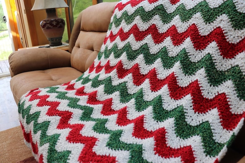 Pu&ograve; includere: Una coperta in crochet chevron di colore rosso, bianco e verde. La coperta &egrave; appoggiata su un divano in pelle marrone.