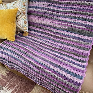 May include: A striped crocheted throw blanket in shades of purple, gray, and pink. The blanket is draped over a brown leather couch.