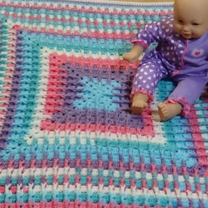 May include: A crocheted baby blanket in shades of pink, blue, purple, and white. The blanket has a square pattern and is made with a soft yarn.