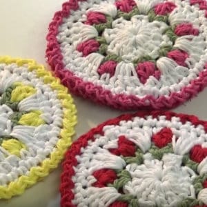 May include: Three handmade crochet coasters in a circular floral design. One coaster has a yellow border, another has a pink border, and the third has a red border. The coasters are white with pink, red, and green accents.