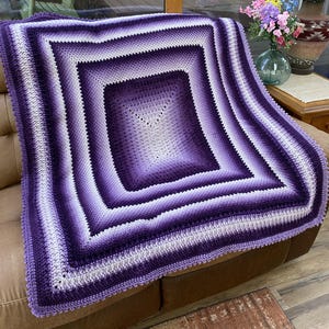 Może przedstawiać: Koc wykonany na szydełku w kolorze fioletowym i białym z kwadratowym wzorem. Koc jest wykonany z włóczki gradientowej, która przechodzi od ciemnego fioletu do jasnego bieli.