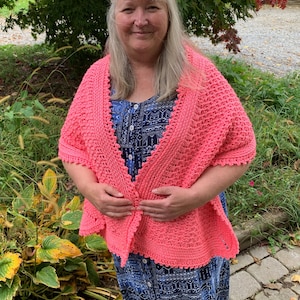 May include: A coral pink crocheted shawl with a scalloped edge. The shawl is worn by a person with long, gray hair.