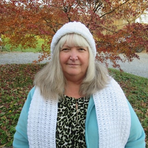 May include: A woman wearing a white crocheted hat and scarf with a blue jacket and a leopard print shirt.