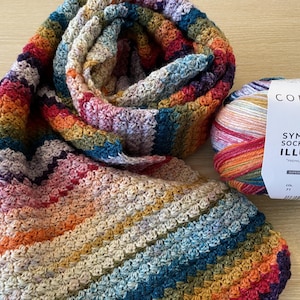 Może przedstawiać: Kolorowy, szydełkowy szalik w odcieniach czerwieni, pomarańczy, żółci, zieleni, niebieskiego i fioletu. Obok znajduje się motek włóczki z napisem "CONCEPT BY KATIA 4 PLY SYMMETRIC SOCKS & MORE ILLUSION".