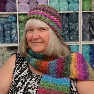 Puede incluir: Una mujer lleva un gorro y una bufanda de ganchillo de colores. El gorro y la bufanda están hechos con una variedad de colores, incluyendo verde, azul, rosa y morado. La mujer está de pie frente a una pared de hilo.