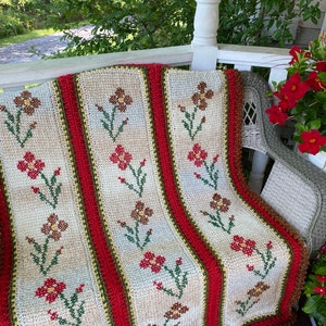 May include: A crocheted throw blanket with a floral pattern in shades of red, brown, and green. The blanket is draped over a white wicker chair on a porch.