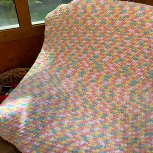 May include: A crocheted baby blanket in a pastel color scheme of pink, blue, yellow, and white. The blanket is made with a soft, fluffy yarn and has a textured, bumpy surface.