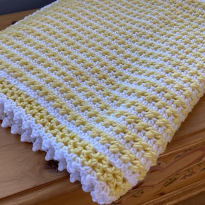 May include: A handmade, crocheted blanket in yellow and white. The blanket features a textured stitch pattern with alternating stripes of yellow and white. The edges are finished with a white, scalloped trim. The blanket is folded on a wooden surface.