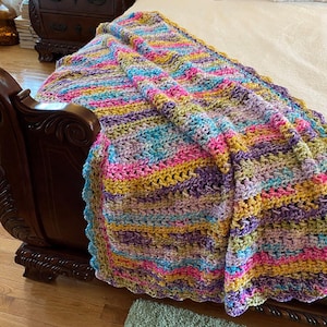 Puede incluir: Una manta de crochet a rayas de colores con un borde festoneado, drapeada sobre una cama con ropa de cama blanca.