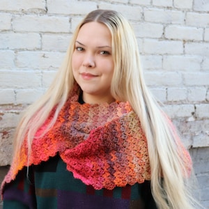 Peut inclure: Un col crocheté coloré aux tons orange, rose et violet. Le col fait main a un bord festonné et se porte autour du cou. L'arrière-plan est un mur de briques blanches.