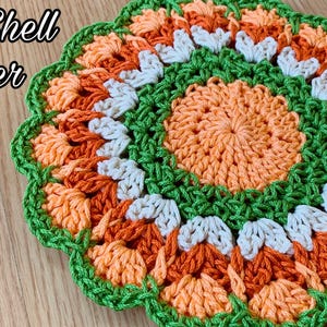 Könnte beinhalten: Ein gehäkelter Topflappen in Form einer Blume mit orangefarbenem, pfirsichfarbenem, weißem und grünem Garn. Der Text "Fancy Shell Potholder" ist im Bild sichtbar.