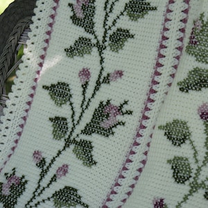 May include: A cream-colored, crocheted blanket with a floral cross-stitch design. The blanket features green leaves and pink flowers, with a scalloped edge and a border of pink triangles. The blanket is draped over a wicker chair.